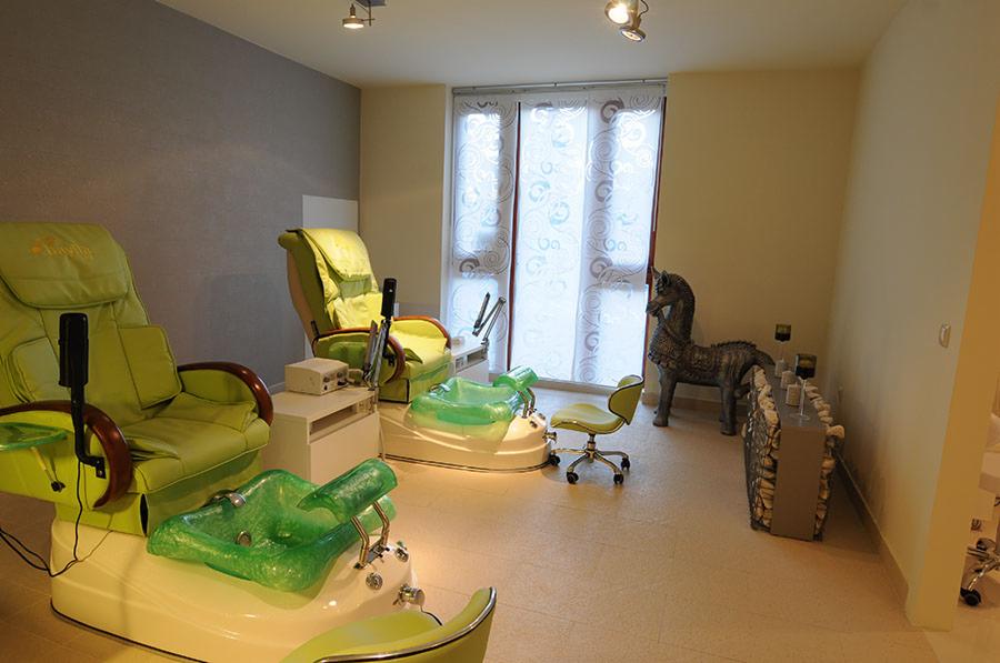 Wnętrze salonu kosmetycznego z dwoma fotelami do pedicure w odcieniach zieleni, z podnóżkami w kształcie muszli i lampami, dekoracyjne zasłony w oknach oraz rzeźba konia na tle ściany.