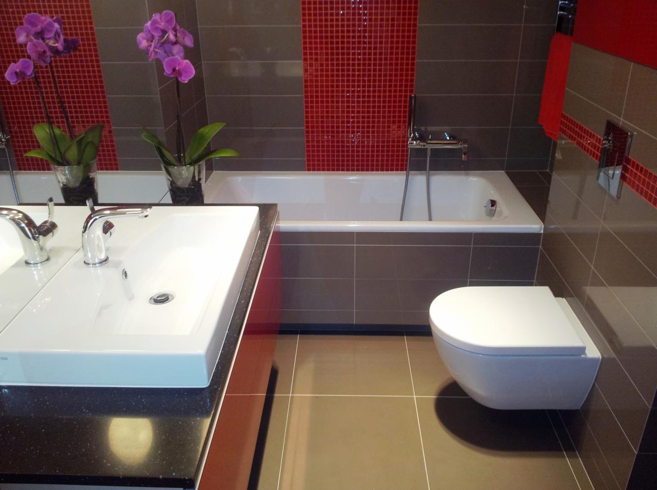 Nowoczesna łazienka z umywalką na czarnym blacie, wolnostojącą toaletą i wanną obłożoną szarą mozaiką z czerwonym akcentem, dopełniona fioletowymi storczykami.