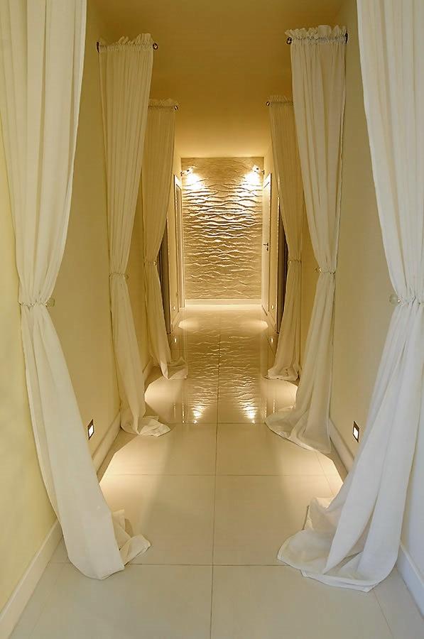 Korytarz z białymi płytkami podłogowymi, dekoracyjną ścianą z efektem fal i białymi zasłonami po bokach, oświetlony punktowymi lampami.