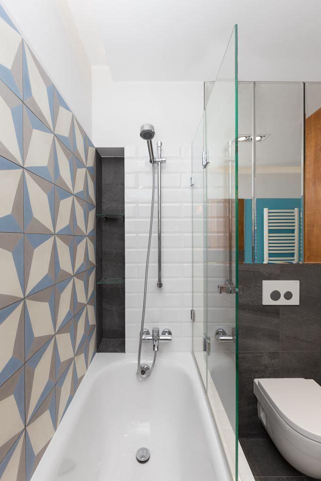 Nowoczesna łazienka z wanną, szklanym parawanem nawannowym, geometrycznymi płytkami ściennymi w odcieniach błękitu i szarości oraz białą armaturą, w tle widoczne lustro i grzejnik.