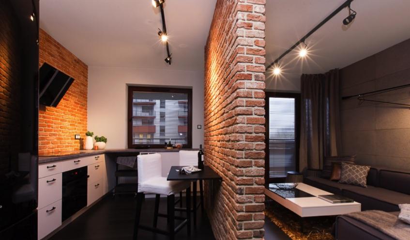 Nowoczesne wnętrze mieszkania z ceglaną ścianą działową, czarnymi lampami na szynoprzewodach, aneksem kuchennym i częścią wypoczynkową z szarą sofą.