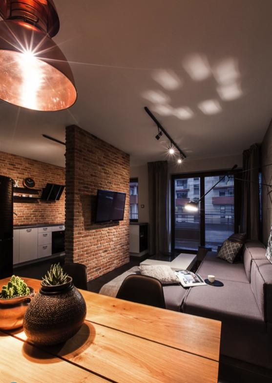Nowoczesne wnętrze mieszkania z ceglaną ścianą działową, oświetleniem szynowym i drewnianym stołem na pierwszym planie.