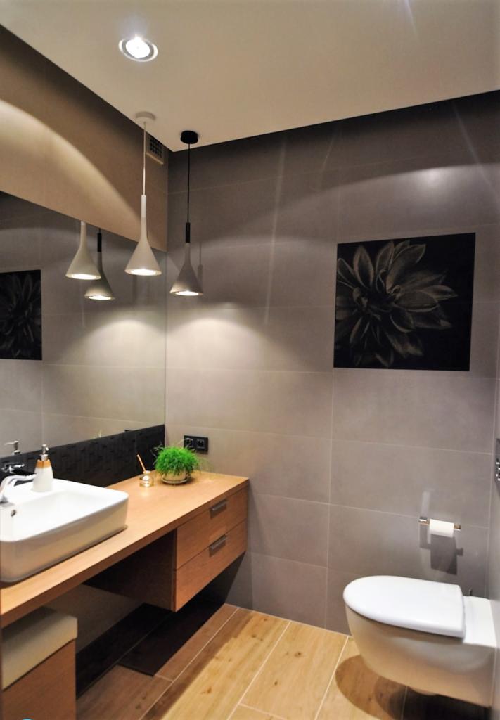 Nowoczesna łazienka z drewnianą podłogą i szafką pod umywalką, szare płytki ścienne, czarno-biały obraz kwiatu i wiszące lampy stożkowe.