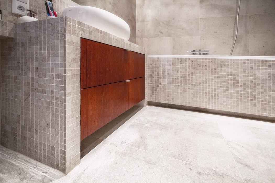 Nowoczesna łazienka wykończona mozaiką w odcieniach szarości, z drewnianą szafką pod umywalką i wanną w tle.