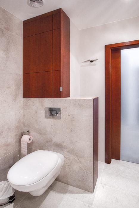 Nowoczesna toaleta podwieszana z szarą marmurową okładziną ścienną i wiszącą szafką z ciemnego drewna, widoczny chromowany przycisk spłuczki i rolka papieru toaletowego.