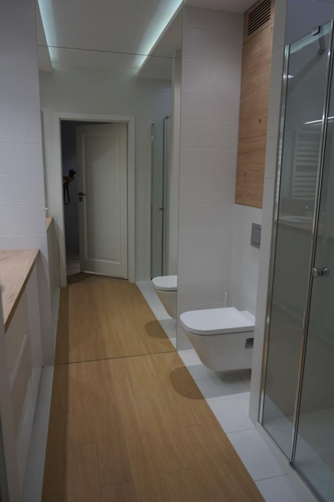 Nowoczesna łazienka z drewnopodobną podłogą, białymi płytkami i minimalistyczną toaletą podwieszaną, widoczna kabina prysznicowa z przezroczystego szkła i drewniana zabudowa nad nią.