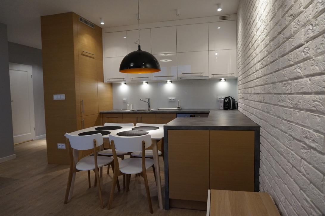 Nowoczesna kuchnia z białymi szafkami, drewnianymi elementami i ścianą z białej cegły, oświetlona punktowymi lampami i wiszącą lampą z czarnym kloszem.