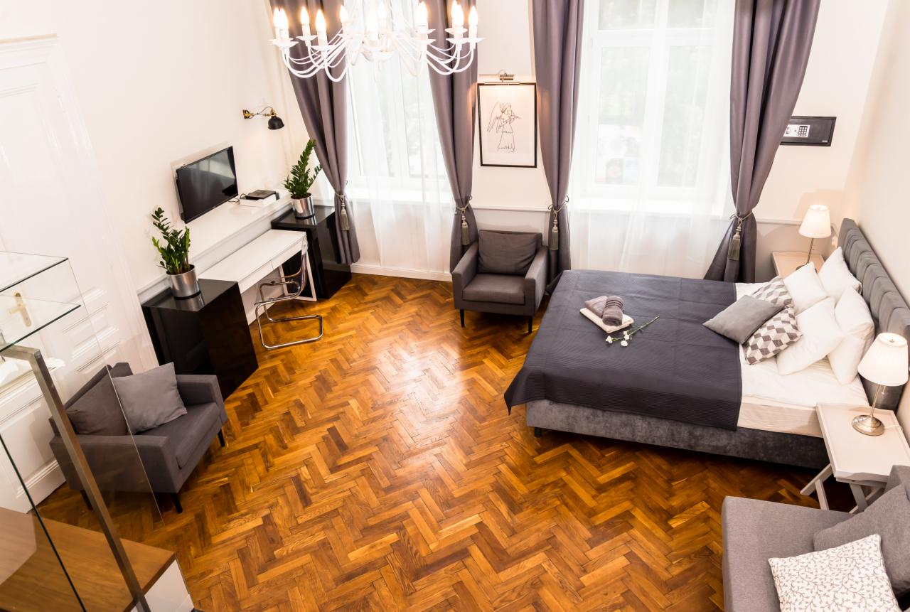 Wysoki kąt widzenia na elegancką sypialnię z parkietem w jodełkę, szarymi zasłonami i meblami, białymi ścianami, żyrandolem i roślinami doniczkowymi.