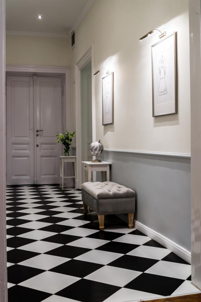 Elegancki korytarz z czarno-białą podłogą w szachownicę, szarym pufem z guzikami, dwoma stolikami z dekoracjami, białymi drzwiami i ścianami z szarą lamperią oraz oświetleniem punktowym nad obrazami.