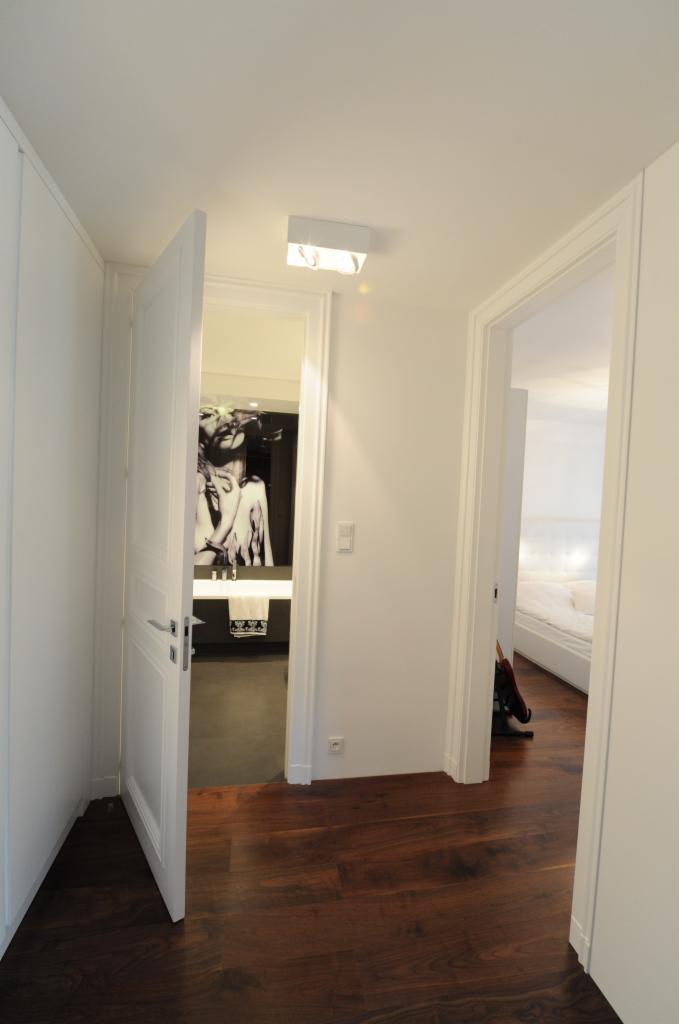 Wnętrze mieszkania z otwartymi drzwiami do łazienki z czarno-białą grafiką na ścianie i widokiem na sypialnię z białym łóżkiem, ciemna drewniana podłoga.