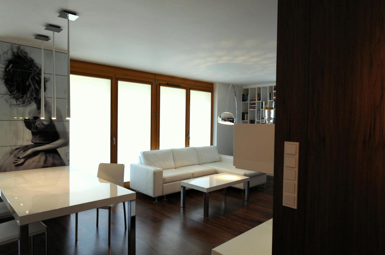 Jasne, przestronne wnętrze z drewnianą podłogą, białym narożnikiem, błyszczącym stołem i krzesłami, oświetleniem punktowym oraz dużym obrazem na ścianie, widok na okna z roletami.