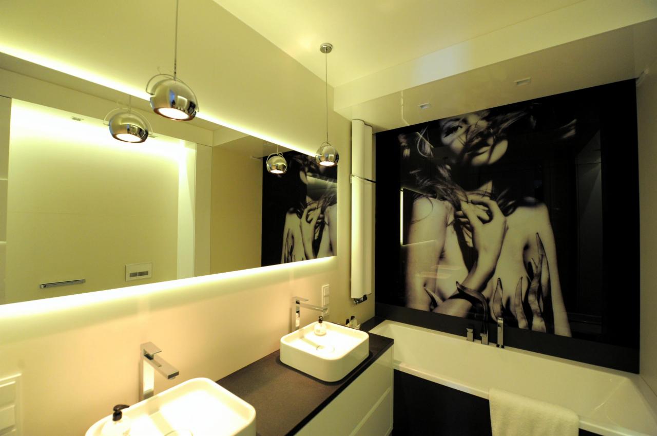 Nowoczesna łazienka z dwoma białymi umywalkami na czarnym blacie, podświetlanym lustrem i grafiką na ścianie nad wanną, oświetlona wiszącymi lampami.