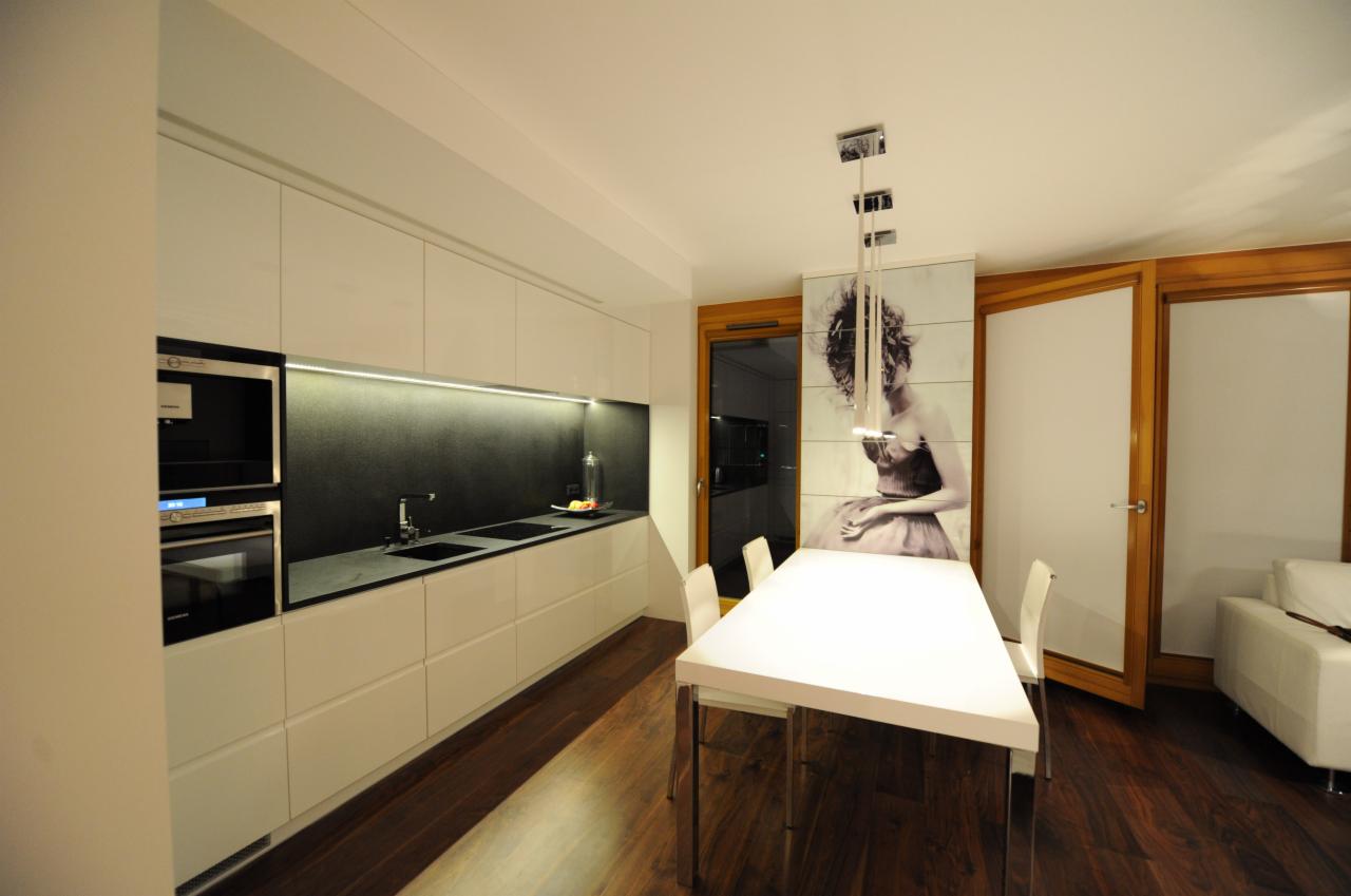 Nowoczesna kuchnia z białymi, lakierowanymi szafkami, czarnym blatem, wbudowanym sprzętem AGD i drewnianą podłogą. Nad stołem minimalistyczna lampa, a na ścianie dekoracyjny panel z portretem kobiety.