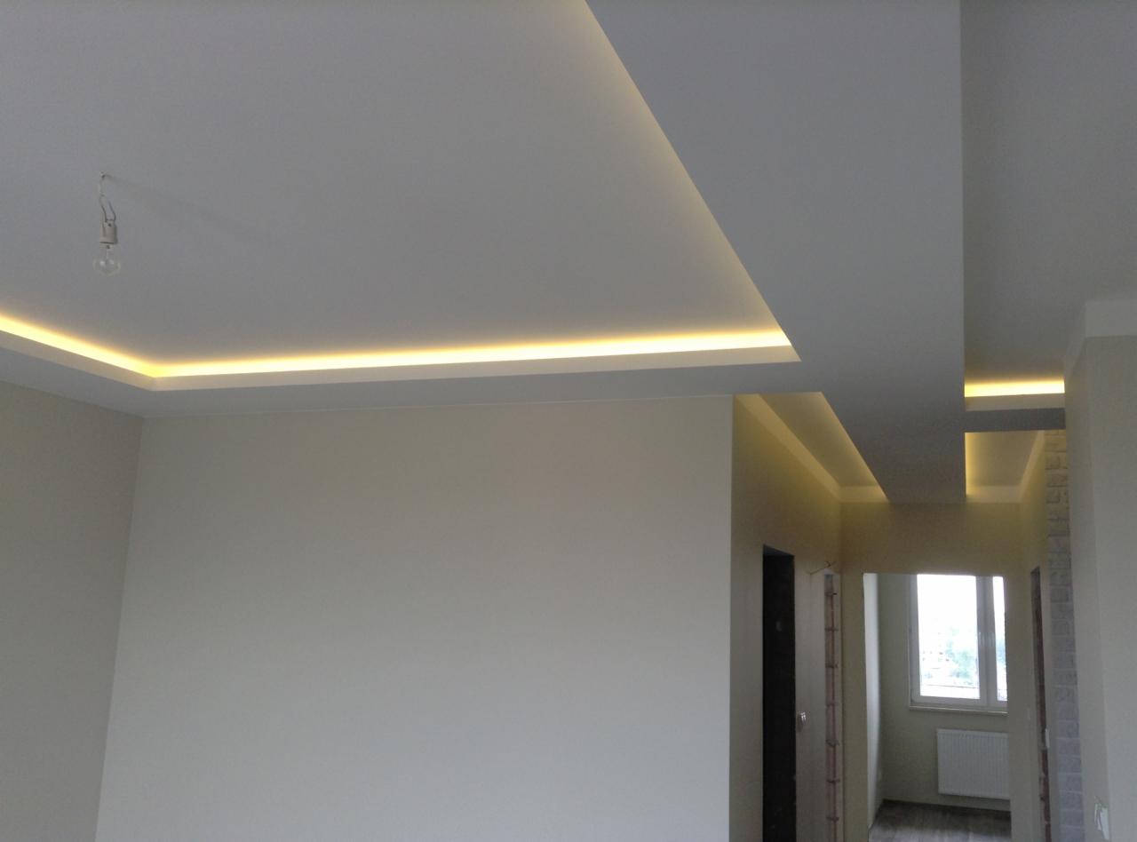 Nowoczesne oświetlenie sufitowe LED w kształcie prostokątów, z ciepłym białym światłem, w jasnym, odświeżonym wnętrzu z widocznym wejściem do kolejnego pomieszczenia.