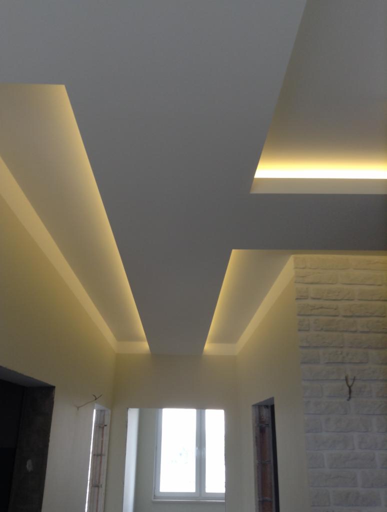 Nowoczesny sufit podwieszany z oświetleniem LED w kształcie geometrycznym, ściana wykończona imitacją białej cegły, widok na okno i przygotowane otwory drzwiowe.