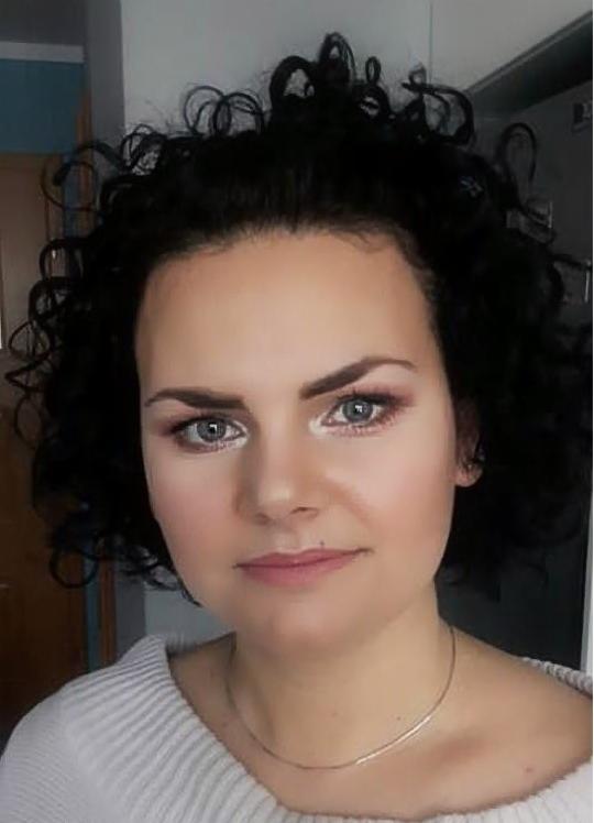 Portret kobiety o krótkich, kręconych, czarnych włosach i delikatnym różowym makijażu oczu i ust, ubrana w biały sweter i cienki złoty łańcuszek.