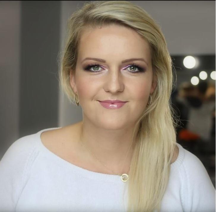 Portret blondynki z delikatnym, różowym makijażem oczu i błyszczącymi ustami, w tle widoczne lampy oświetleniowe.