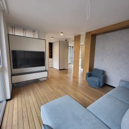 TKACH MONTAGE SERVICE Sergii Tkach - Nowoczesny salon po remoncie: drewniana podłoga, szara sofa, telewizor na ścianie z szafką RTV. Minimalistyczny design z elementami drewna i jasnymi ścianami.