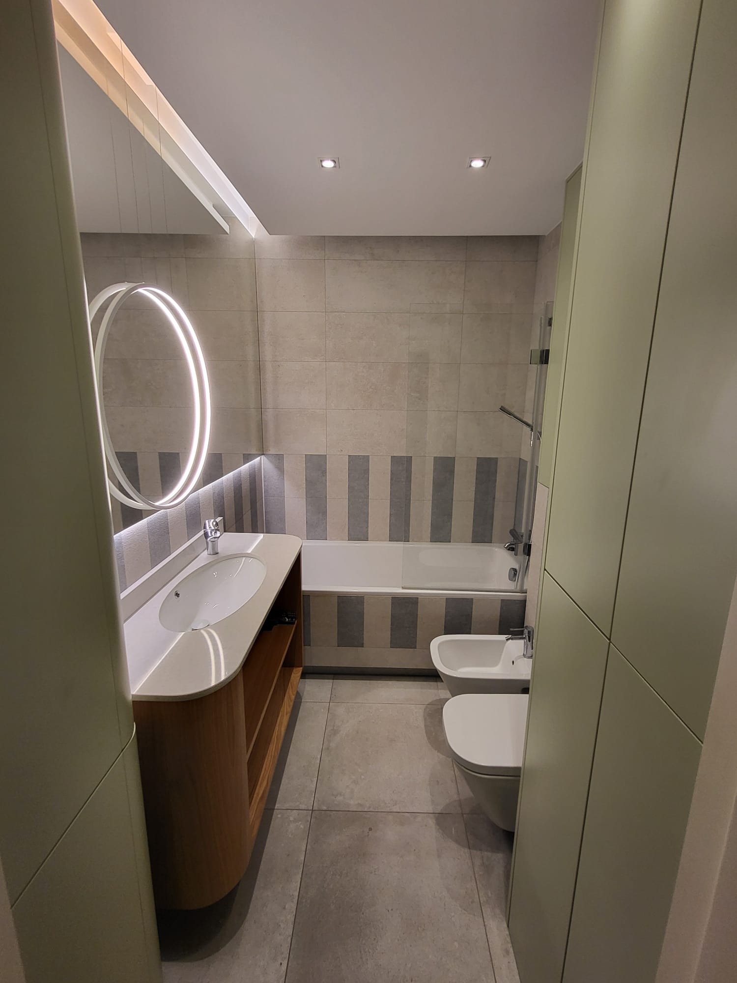 Nowoczesna łazienka z okrągłym lustrem podświetlanym LED, umywalką na drewnianej szafce, wanną w pasy i bidetem. Minimalistyczny design, stonowana kolorystyka.