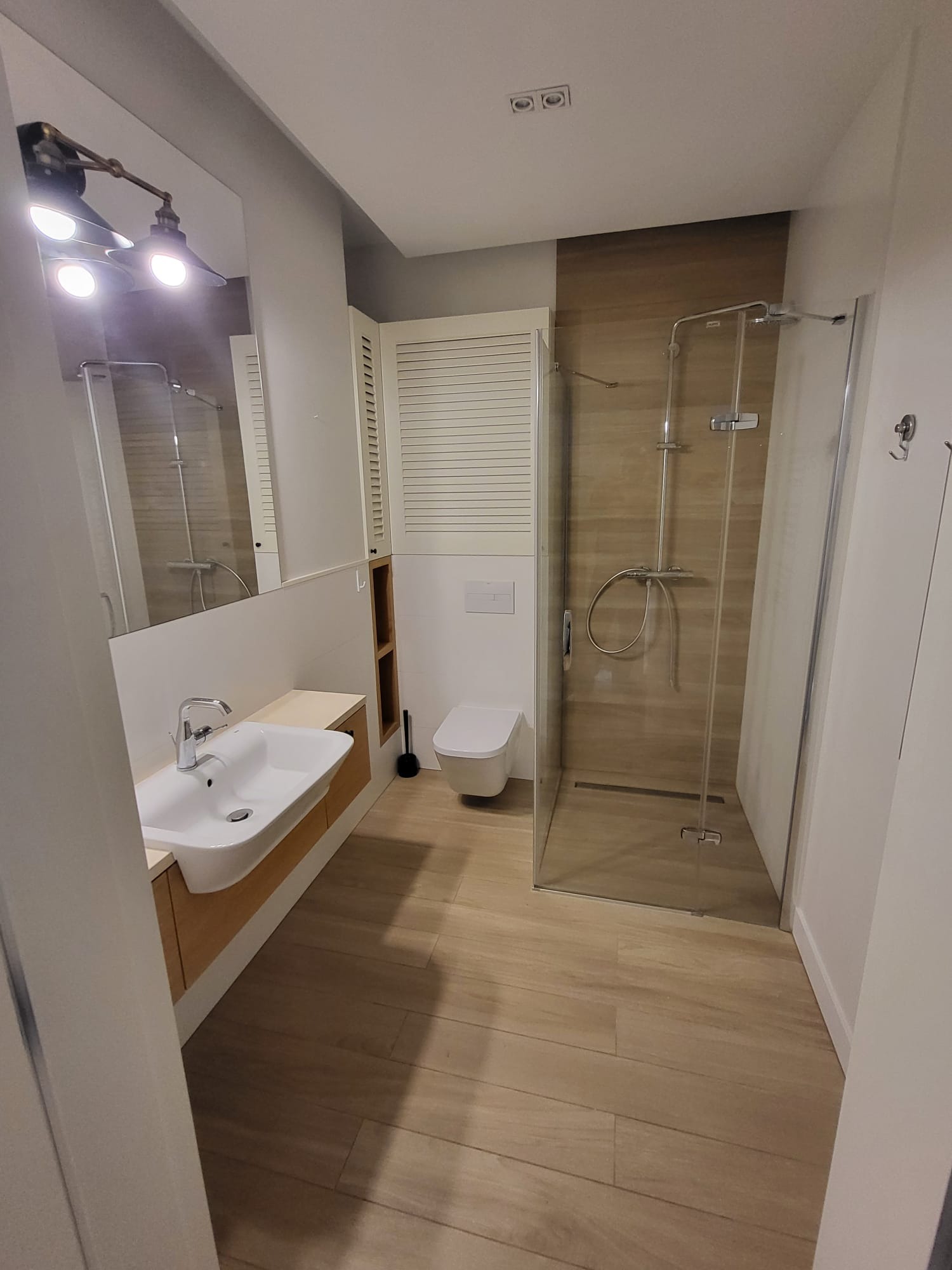 Jasna łazienka z kabiną prysznicową typu walk-in, umywalką nablatową i drewnopodobnymi płytkami. Widoczne oświetlenie w stylu retro nad lustrem.