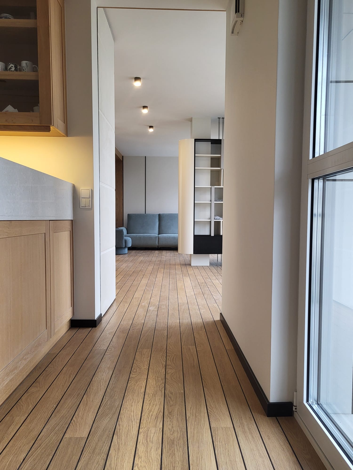 Jasne drewniane panele podłogowe w nowoczesnym wnętrzu. Widok na salon z szarą sofą i regałem. Minimalistyczny design, neutralna kolorystyka, duże okno z prawej strony.