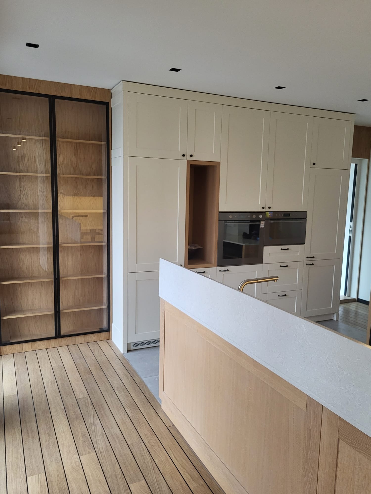 Jasna kuchnia z drewnianą podłogą i zabudową: szafki kremowe, przeszklona witryna z czarnymi ramami, wyspa z białym blatem. Minimalistyczne oświetlenie punktowe.