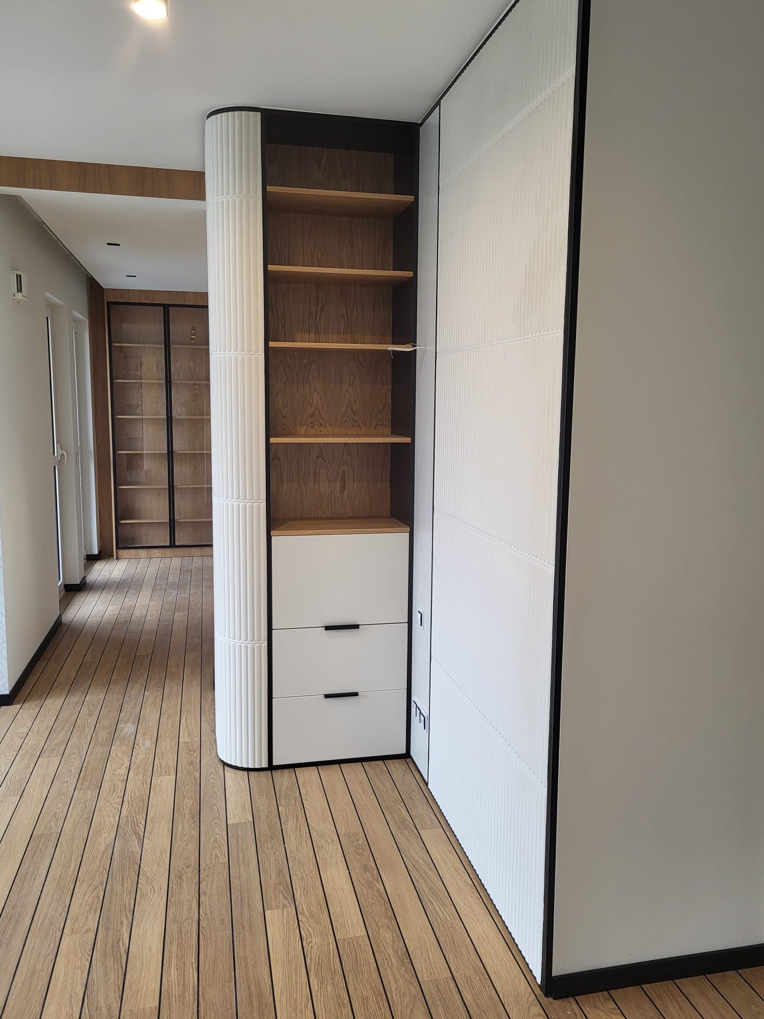 Nowoczesne wykończenie wnętrza: drewniana podłoga, zabudowa z półkami i szufladami, biała ściana z dekoracyjnymi panelami. Minimalistyczny design.