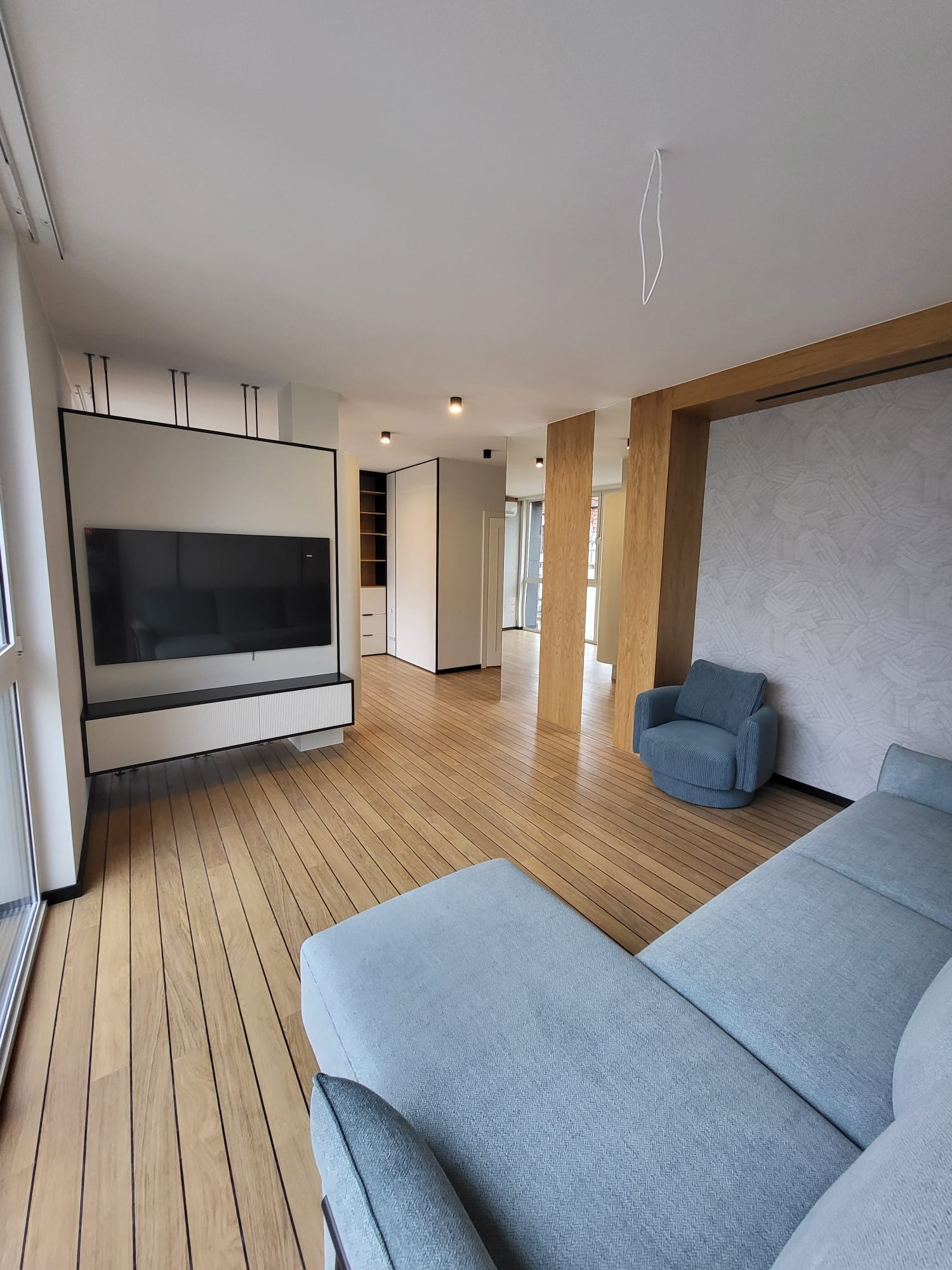 Nowoczesny salon po remoncie: drewniana podłoga, szara sofa, telewizor na ścianie z szafką RTV. Minimalistyczny design z elementami drewna i jasnymi ścianami.