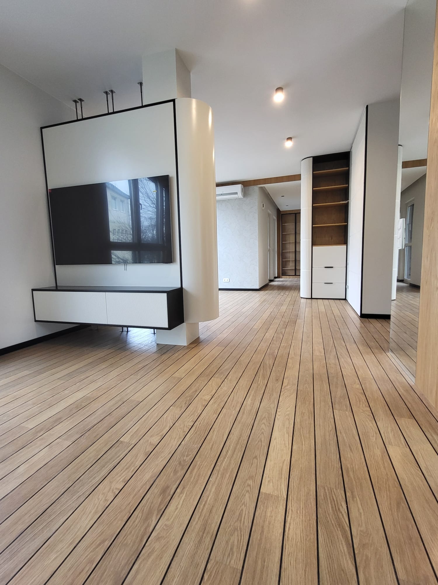 Nowoczesne wykończenie wnętrza: drewniana podłoga z wyraźnym rysunkiem słojów, minimalistyczna zabudowa TV z szafką RTV i kolumną, widok na dalszą część mieszkania.
