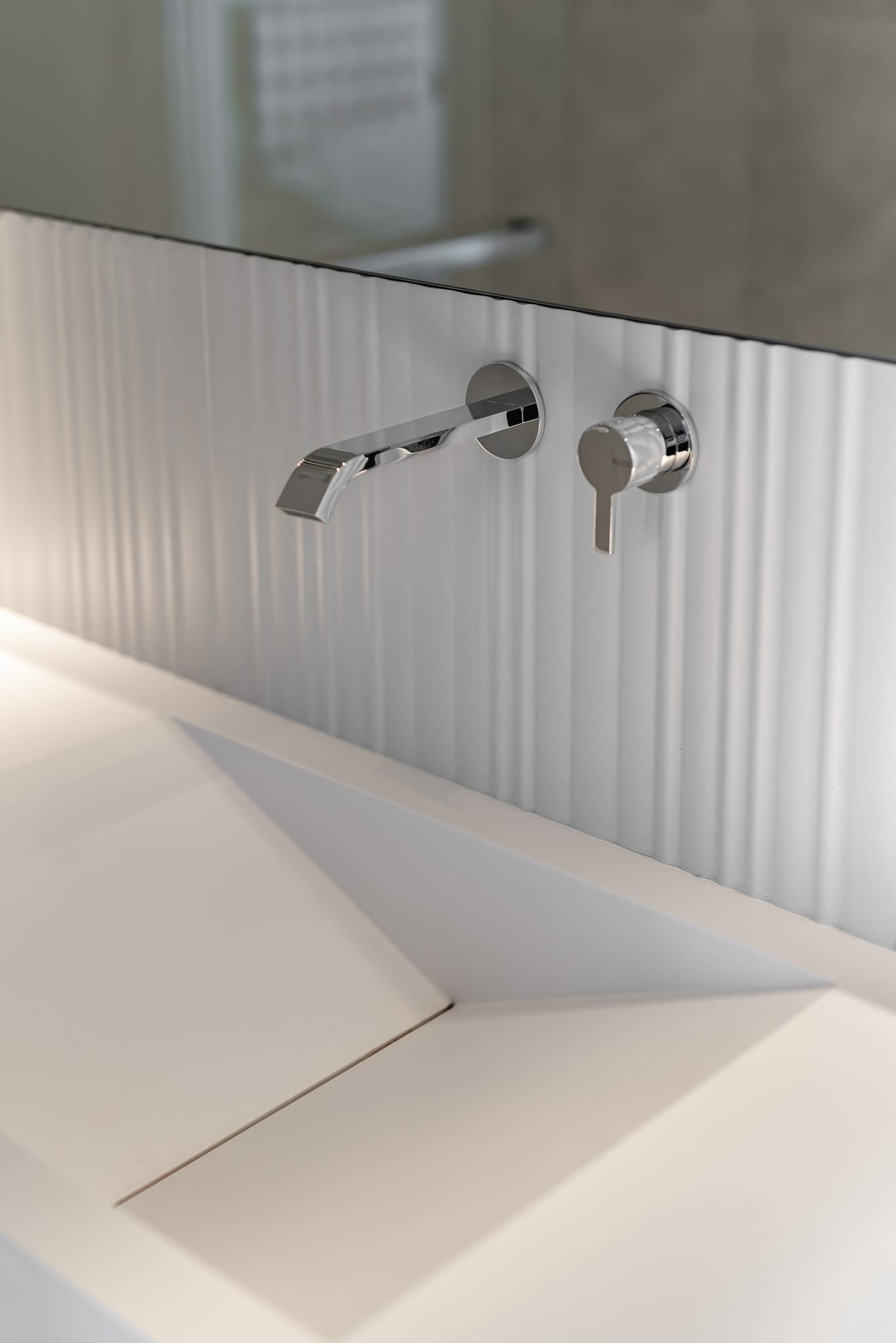 Minimalistyczna umywalka z chromowaną armaturą na tle białej, teksturowanej ściany w nowoczesnej łazience. Geometryczny kształt umywalki, subtelne oświetlenie.