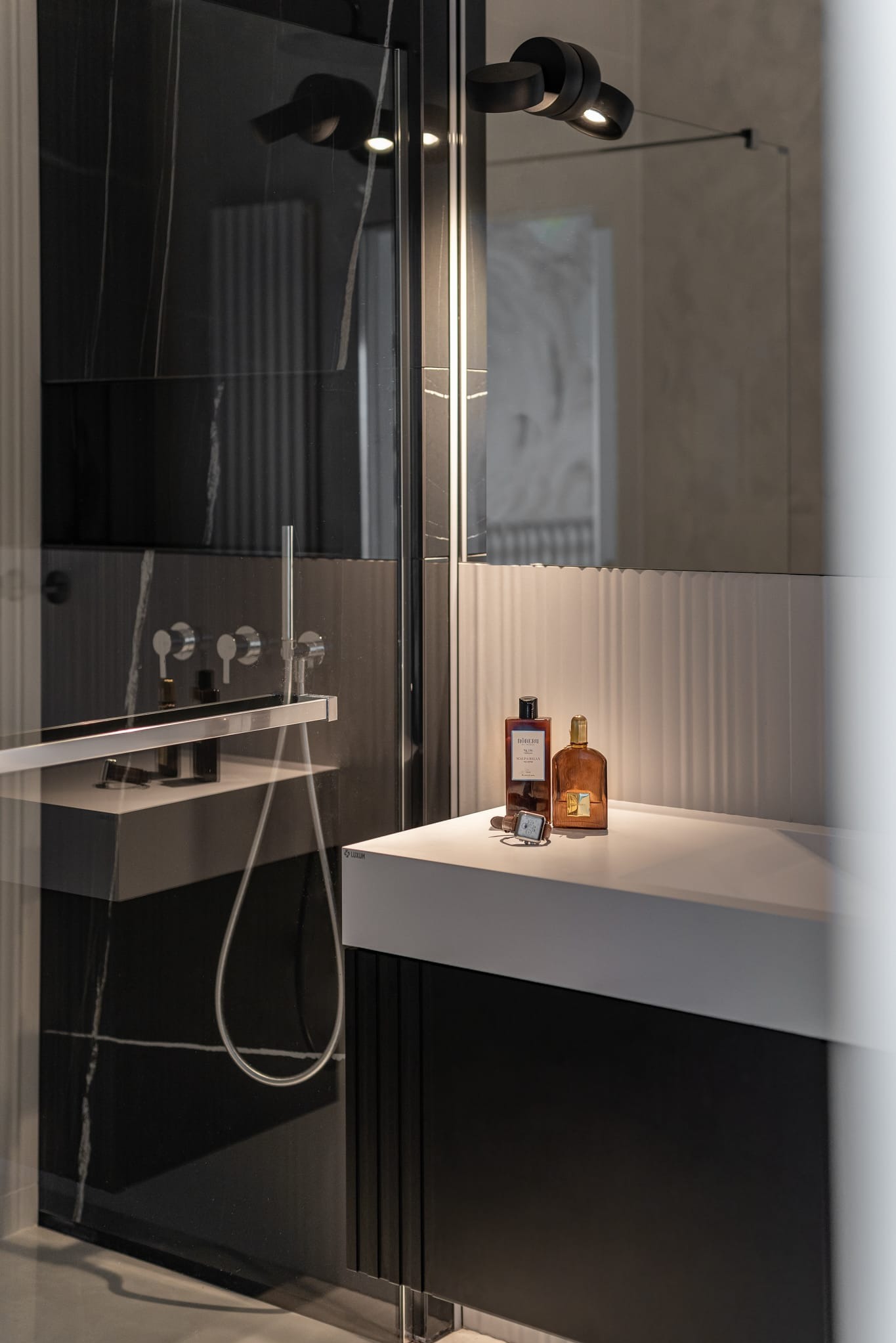 Nowoczesna łazienka z czarnymi płytkami imitującymi marmur, szklaną kabiną prysznicową i białym blatem z kosmetykami. Minimalistyczne oświetlenie punktowe.