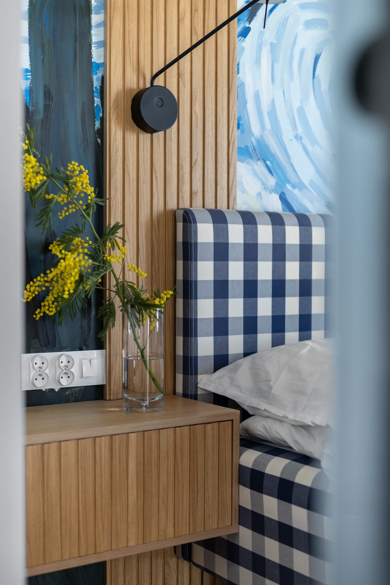 Stylowa sypialnia: drewniana szafka nocna z gniazdkami, wazon z mimozą, zagłówek w niebiesko-białą kratę, lampka i abstrakcyjne obrazy na ścianie.
