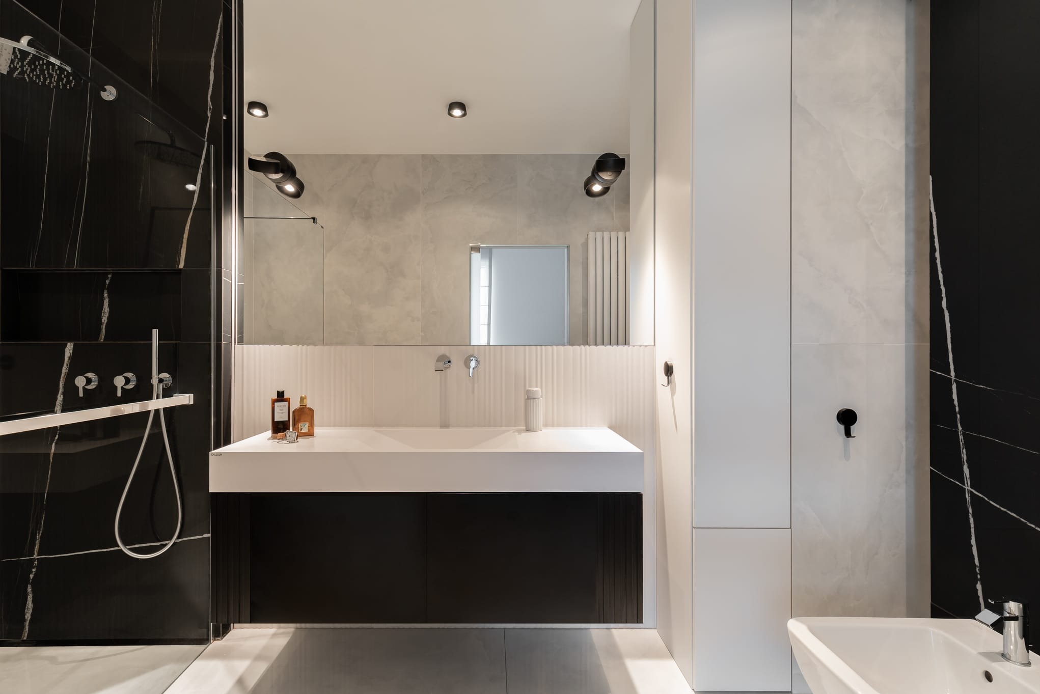 Nowoczesna łazienka z czarno-białymi płytkami imitującymi marmur, minimalistyczną umywalką i szafką. Widoczne oświetlenie punktowe i akcesoria łazienkowe.