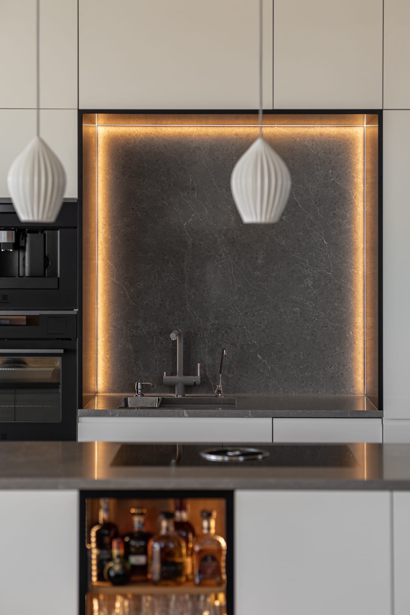 Nowoczesna kuchnia z szarym marmurem, podświetleniem LED i minimalistycznymi lampami wiszącymi. W tle zabudowa z piekarnikiem i ekspresem do kawy.