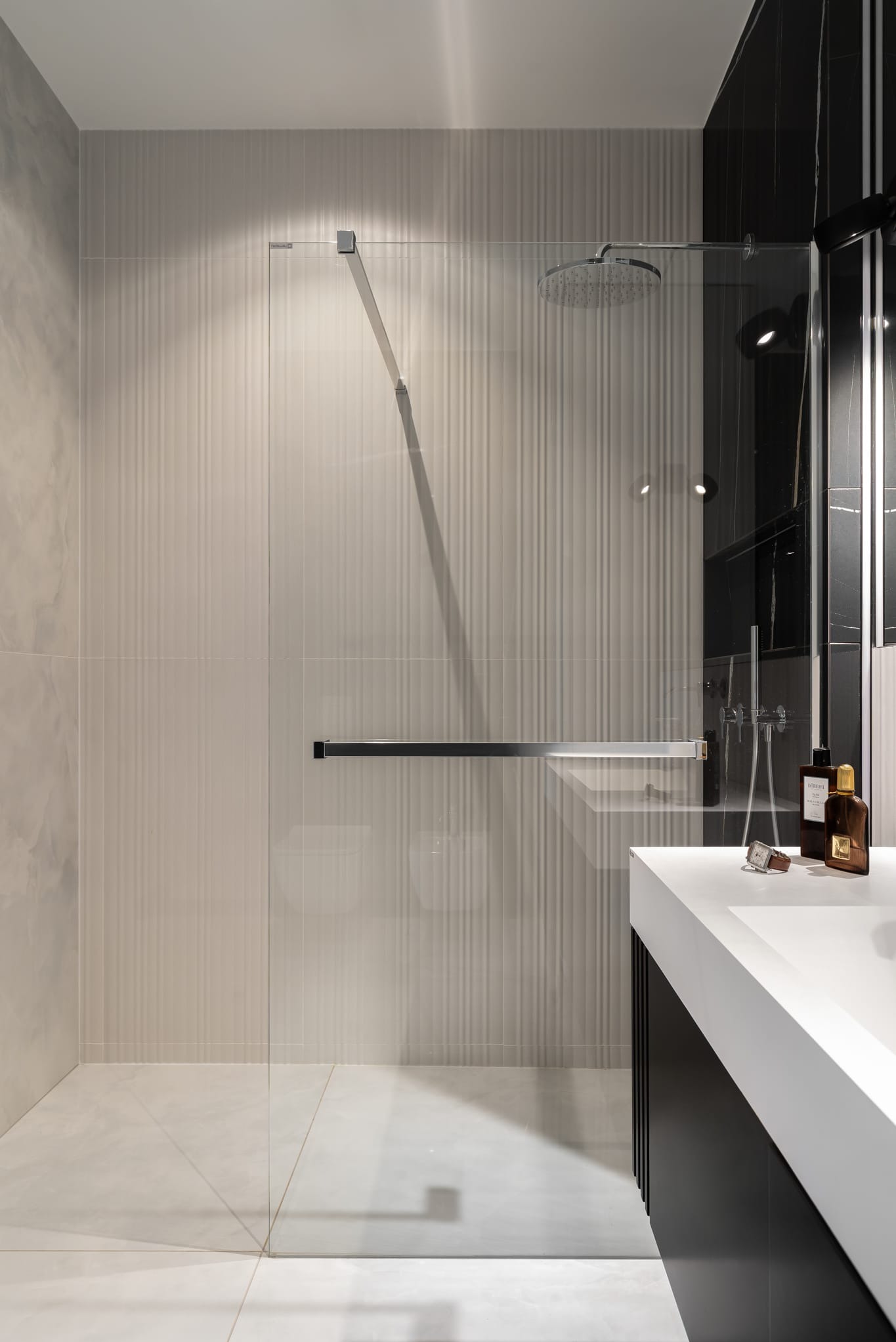 Nowoczesna kabina prysznicowa ze szklaną ścianką, chromowanym uchwytem i deszczownicą. Beżowe płytki z delikatnym wzorem, biała podłoga i czarno-biała umywalka.