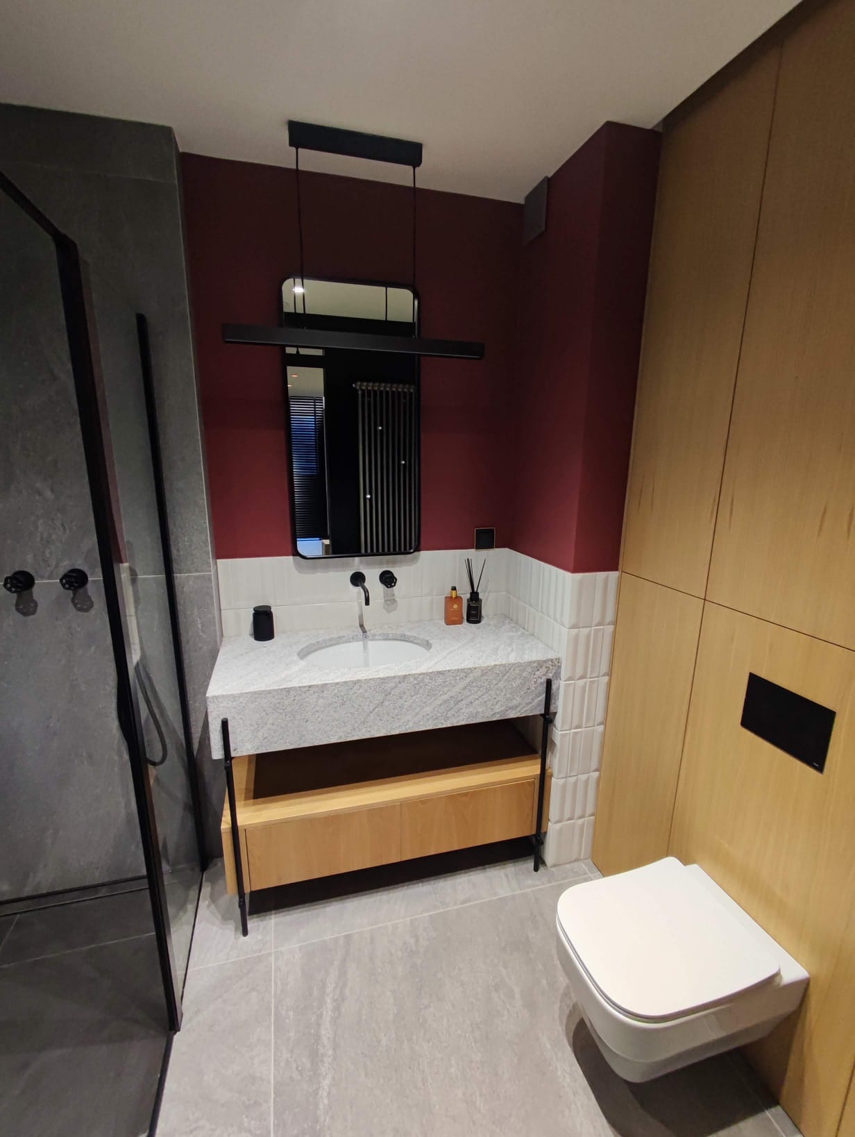 Nowoczesna łazienka z umywalką na blacie z szarego kamienia, wiszącą toaletą i kabiną prysznicową. Ciemnoczerwona ściana i drewniane wykończenia dodają elegancji.
