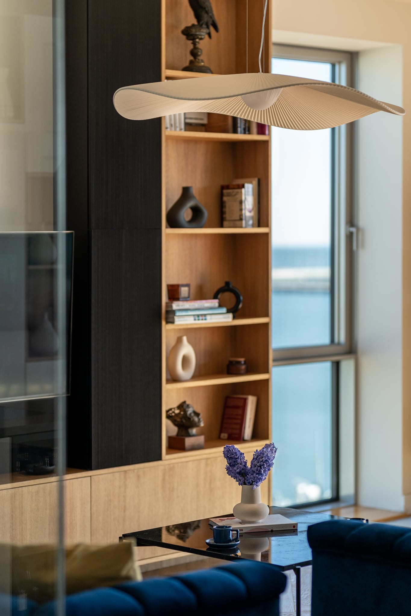 Nowoczesny salon z drewnianą zabudową, półkami z dekoracjami i designerską lampą. Widok na okno z widokiem na morze. Minimalistyczny styl wnętrza.