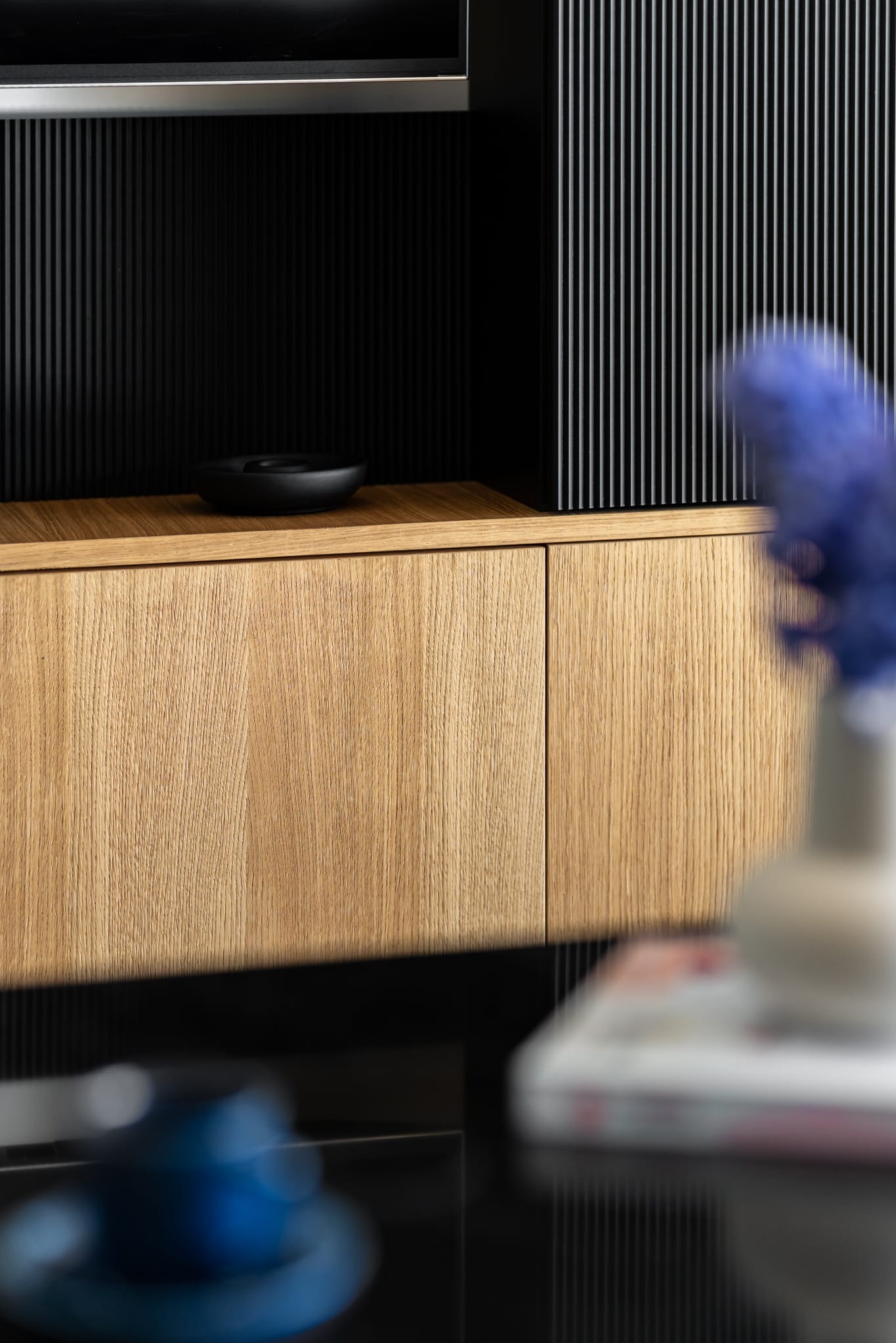 Detal wykończenia wnętrza: drewniana szafka z czarnym, pionowo żebrowanym panelem. Na blacie czarna misa. W tle wazon z niebieskimi kwiatami.