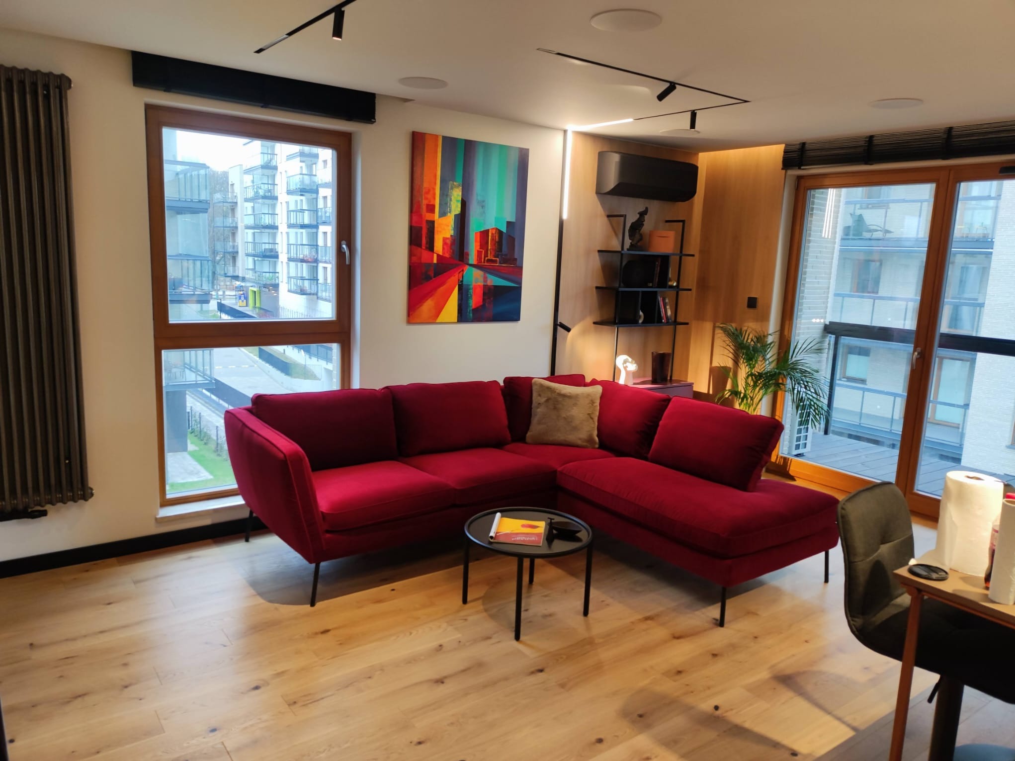 Nowoczesny salon z czerwonym narożnikiem, drewnianą podłogą i abstrakcyjnym obrazem. Widok na budynki przez okno. Minimalistyczne oświetlenie.