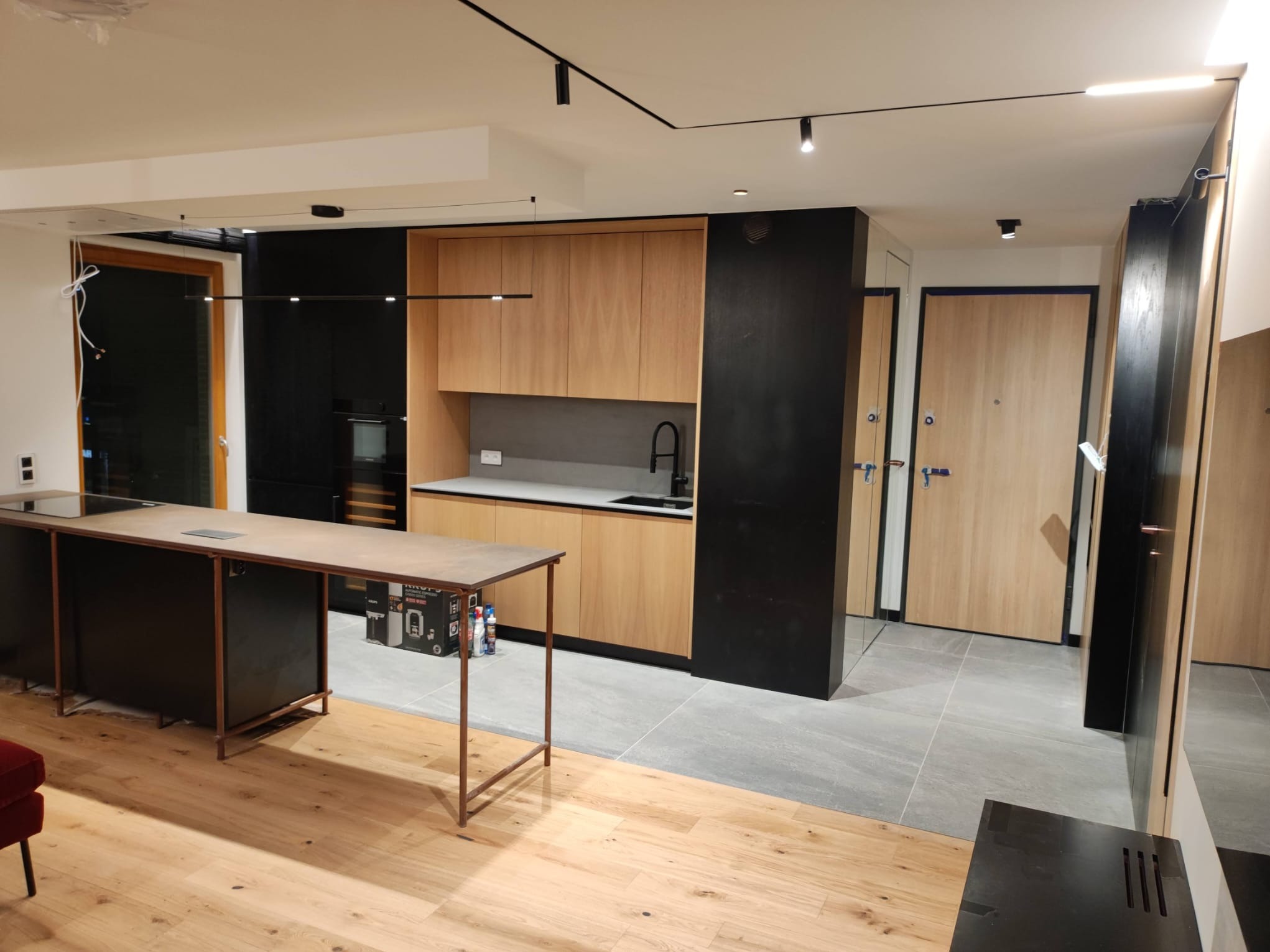 Nowoczesna kuchnia z wyspą w Gdańsku. Jasne drewno szafek kontrastuje z czarnymi elementami. Widoczne oświetlenie punktowe i minimalistyczny design.