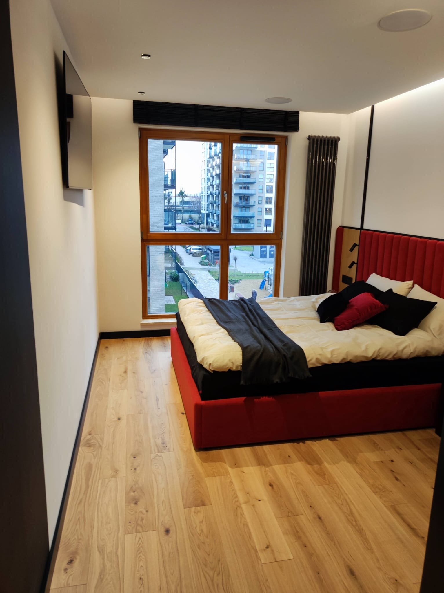 Nowoczesna sypialnia z czerwonym łóżkiem i drewnianą podłogą. Widok na osiedle przez okno. Minimalistyczny design, czarne akcenty i wbudowane głośniki sufitowe.