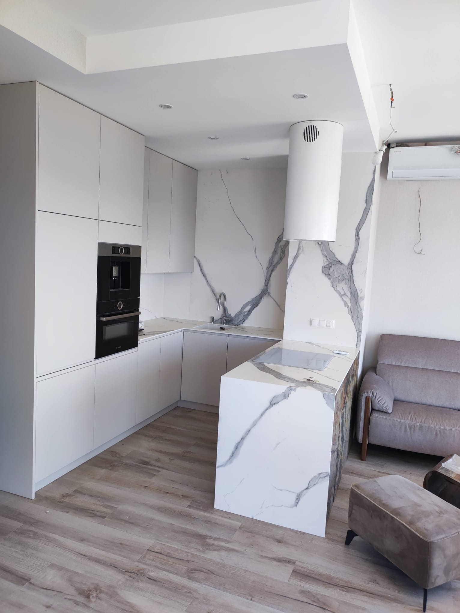 Nowoczesna kuchnia w bieli z marmurowym akcentem na ścianie i wyspie. Minimalistyczne szafki, zabudowany sprzęt AGD i drewnopodobna podłoga tworzą spójną całość.