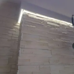 Ściana wyłożona dekoracyjnym kamieniem z podświetleniem LED wzdłuż sufitu, widoczny fragment instalacji elektrycznej z wiszącą lampą bez klosza.