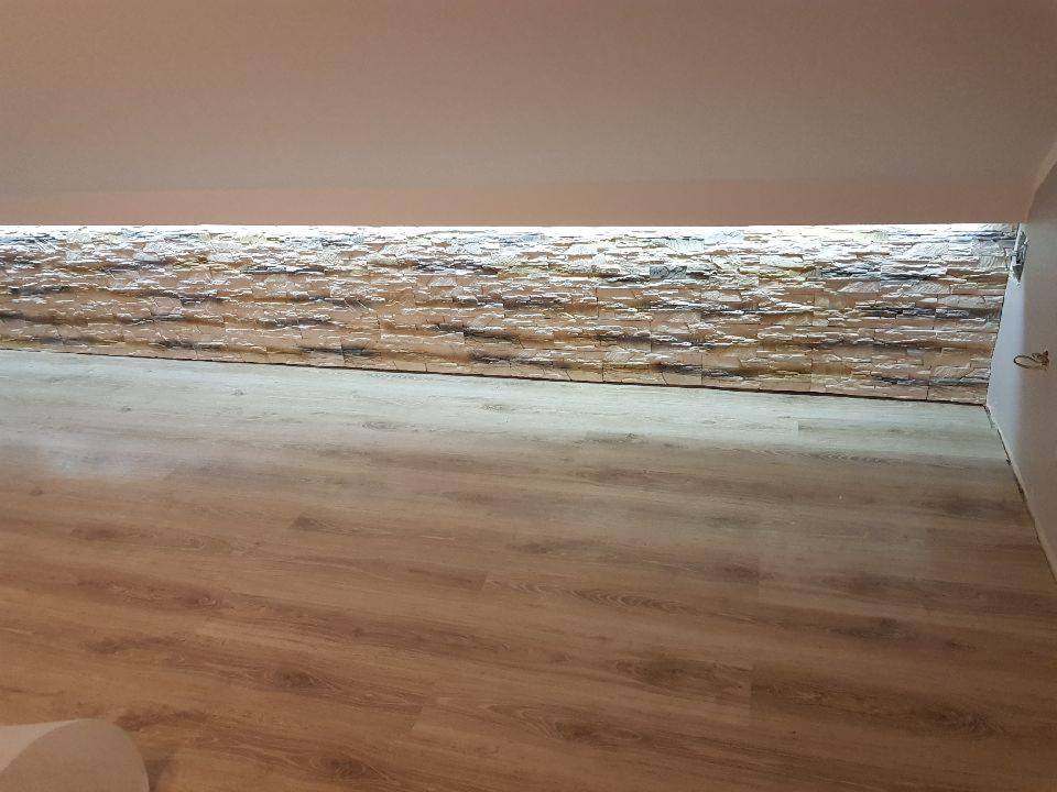 Ściana wykończona dekoracyjną okładziną kamienną z podświetleniem LED, podłoga z paneli drewnopodobnych w pomieszczeniu ze skośnym sufitem.