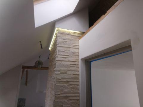 Ściana wykończona kamieniem dekoracyjnym w jasnych odcieniach, z subtelnym oświetleniem LED na krawędzi, widoczna w trakcie prac wykończeniowych na poddaszu, z fragmentem surowego drewna...