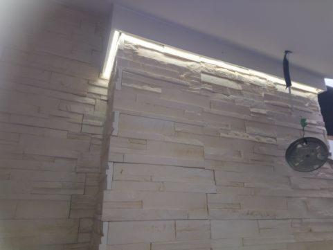 Ściana wyłożona dekoracyjnym kamieniem z podświetleniem LED wzdłuż sufitu, widoczny fragment instalacji elektrycznej z wiszącą lampą bez klosza.