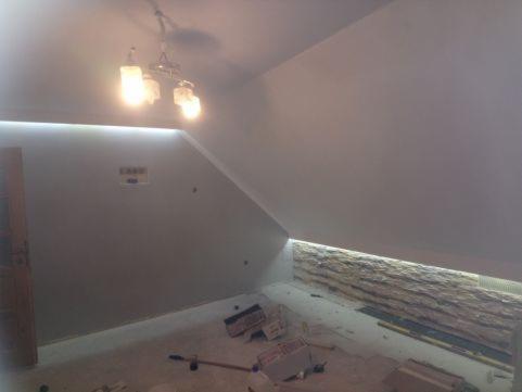 Pokój na poddaszu w trakcie prac wykończeniowych: ściany pomalowane na szaro, widoczna listwa oświetleniowa LED przy suficie, fragment dekoracyjnej ściany z kamienia naturalnego pod oknem,...
