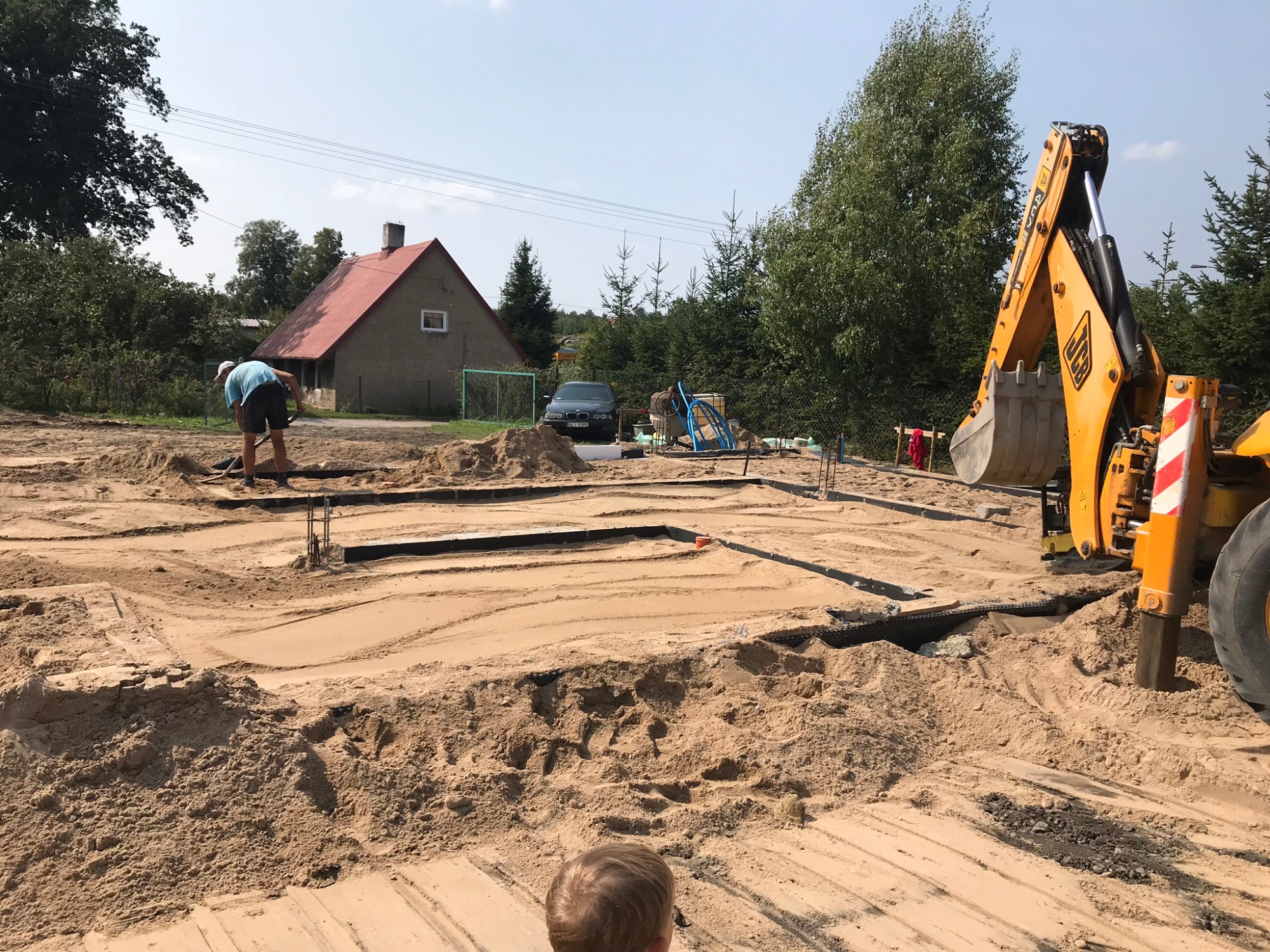 Przygotowanie terenu pod budowę domu jednorodzinnego: wykopane fundamenty, żółta koparka JCB w tle, pracownik wyrównujący teren łopatą, widoczny dom z czerwonym dachem i auto osobowe.