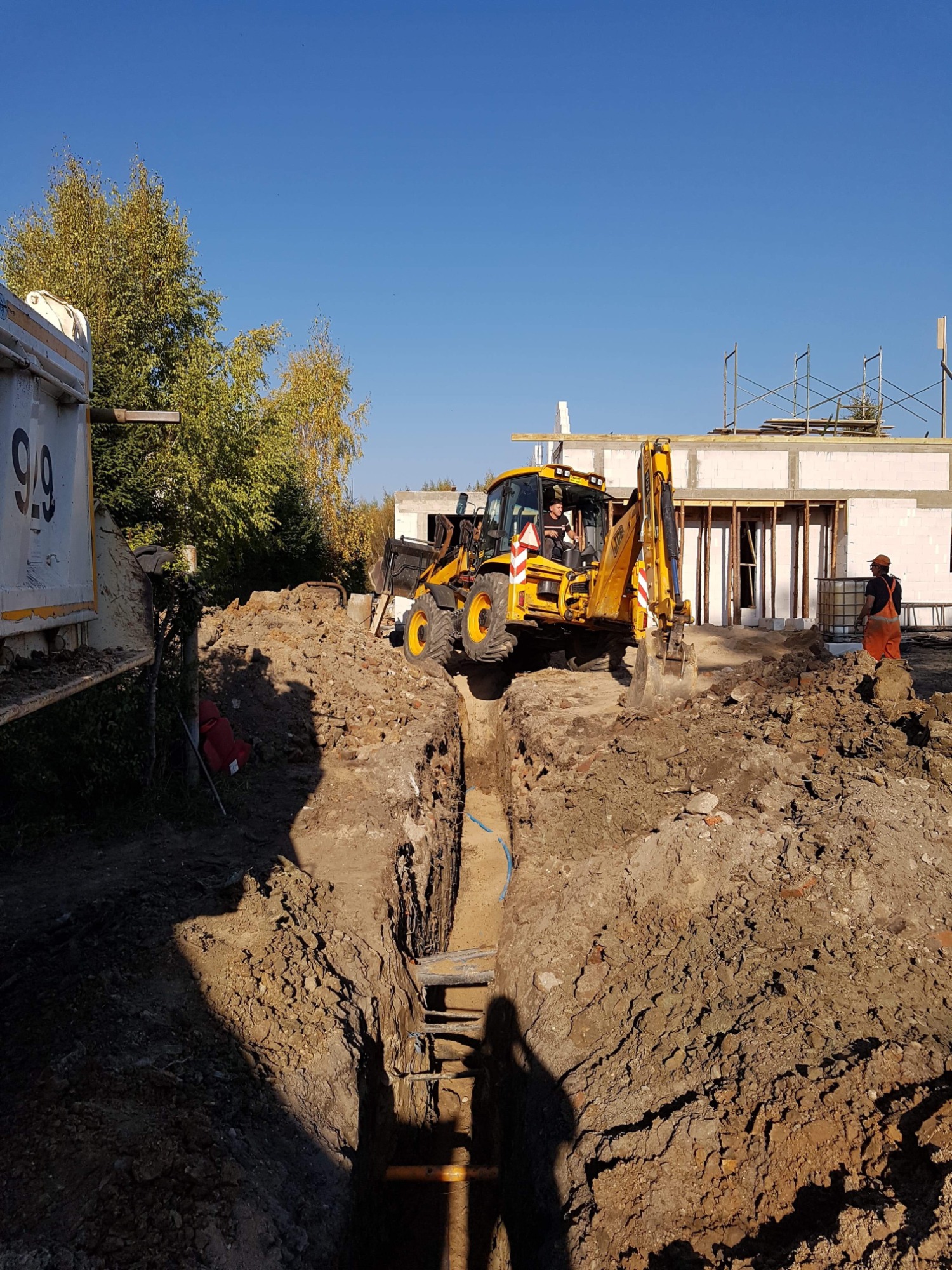 Żółta koparko-ładowarka JCB nad wykopem z widocznymi rurami, w tle budynek w trakcie budowy i pracownik w pomarańczowym kombinezonie.