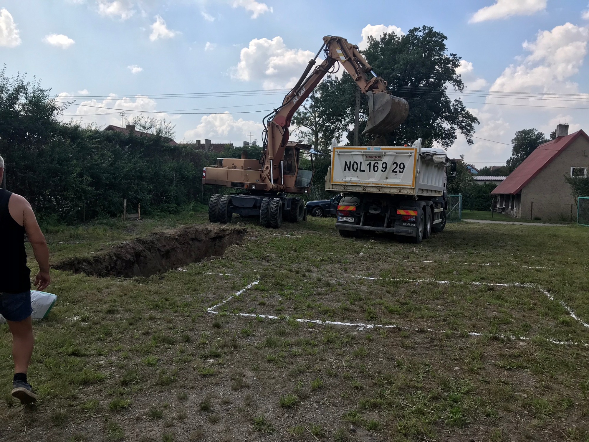 Brązowy koparko-ładowarka Atlas nabiera urobek do białej wywrotki MAN z numerem rejestracyjnym NOL 16929, obok wykopany rów i wyznaczone białą linią obrysy fundamentów, w tle zabudowania.